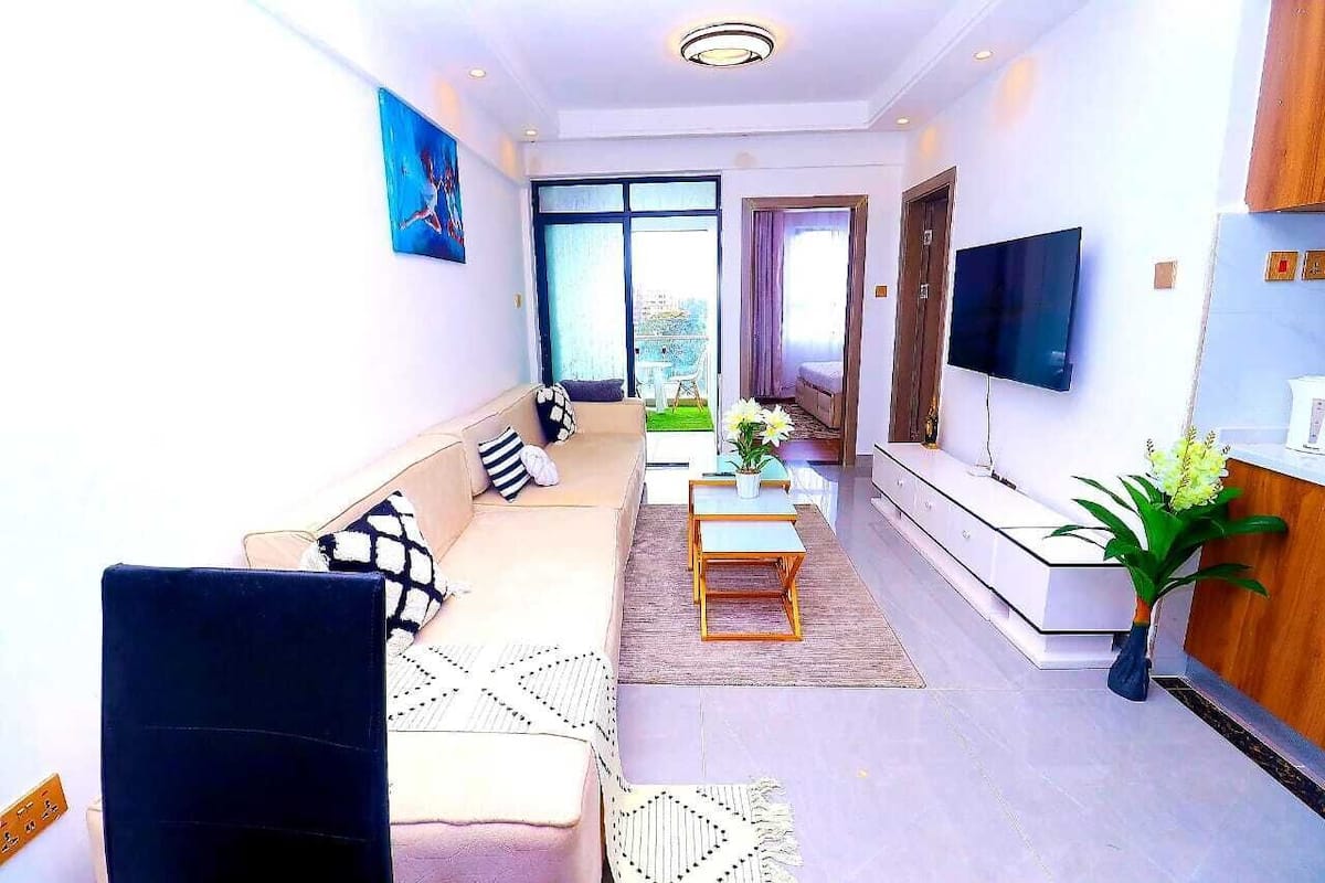 A modern living room with a sofa, coffee table, and TV. There is a balcony with a view.