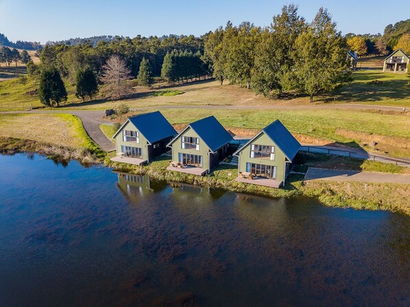 A row of three green houses with dark roofs, situated by a calm body of water, surrounded by a grassy landscape and trees. - Springholm Cottages (Nottingham Road)