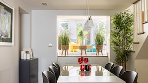 A dining area with a white table, black chairs, and red wine glasses. There are potted plants on the windowsill and a large plant beside the table.
