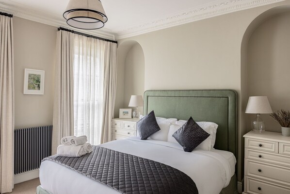 A neatly arranged hotel room with a large bed, two bedside tables with lamps, a radiator, and a window with curtains.