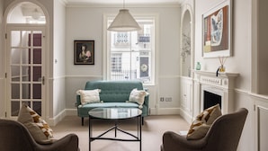 A living room with a teal sofa, two armchairs, a glass coffee table, a white fireplace, and a large window with a view of the city.