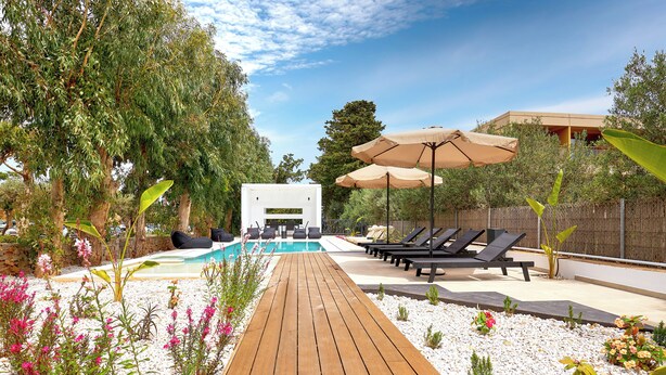 Una terrassa de fusta que condueix a una zona de piscina amb gandules i ombrelles, envoltada de vegetació i un edifici blanc.