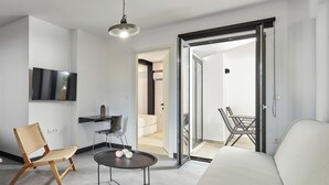 Un saló modern amb un sofà, una cadira de vimetla, una taula petita amb objectes decoratius, un televisor de pantalla plana fixat a la paret i una porta corredissa de vidre que dona a una altra estança.