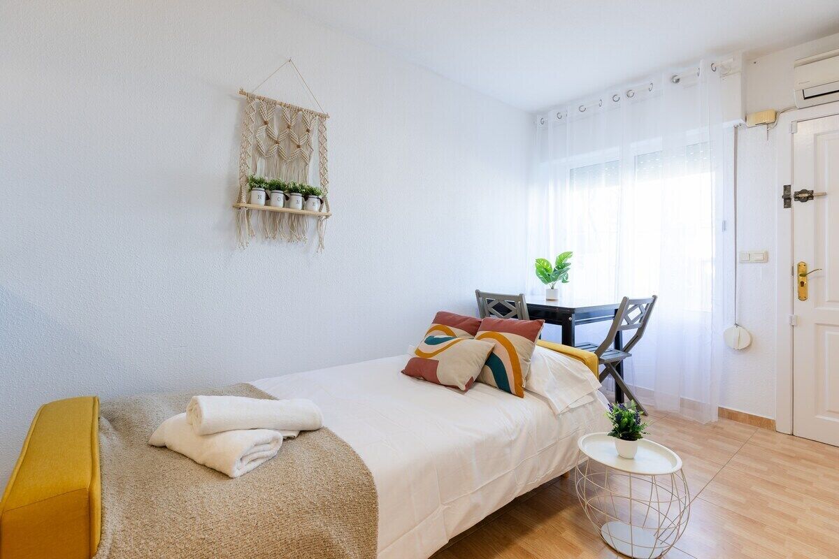 A bedroom with a bed, a small table with chairs, a macrame wall hanging, and a potted plant.