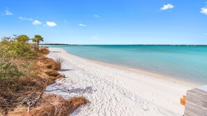 Ein Strand mit weißem Sand, klarem türkisblauem Wasser und vereinzelter Vegetation.