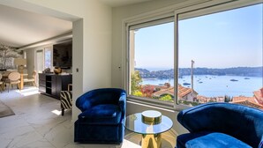 A spacious living room with a large window offering a view of a bay with boats. There are two blue armchairs and a small glass table. - VILLA  BELLE VUE VI4394 By Riviera Holiday Homes (Villefranche-sur-Mer)