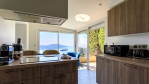 A modern kitchen with a view of the sea, a dining area with a blue chair, and a large window. - VILLA  BELLE VUE VI4394 By Riviera Holiday Homes (Villefranche-sur-Mer)