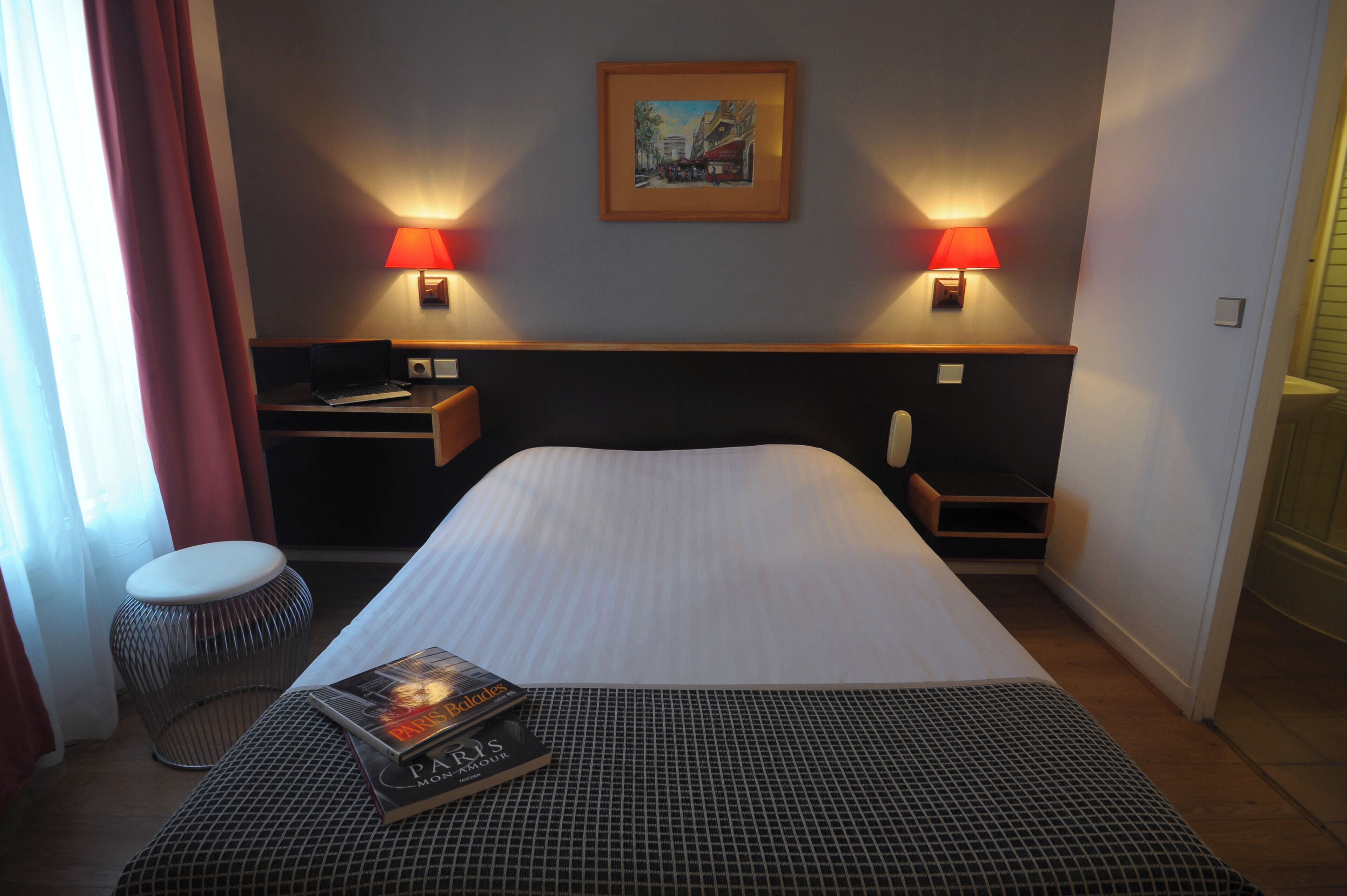 A hotel room with a bed, two bedside lamps, a desk with a laptop, a chair, a small round stool, and a framed picture on the wall.