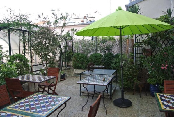 Un patio al aire libre con mesas y sillas, rodeado de follaje y un paraguas verde.