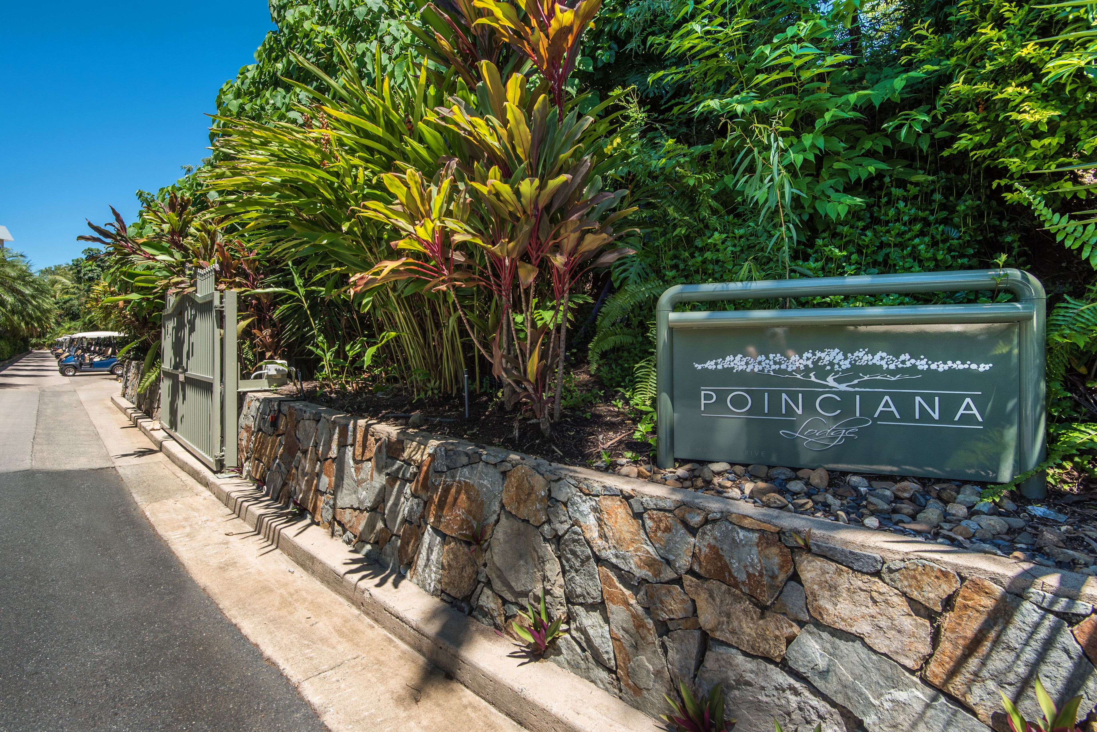 A sign for Poinciana Lodge with a stone wall and lush greenery.