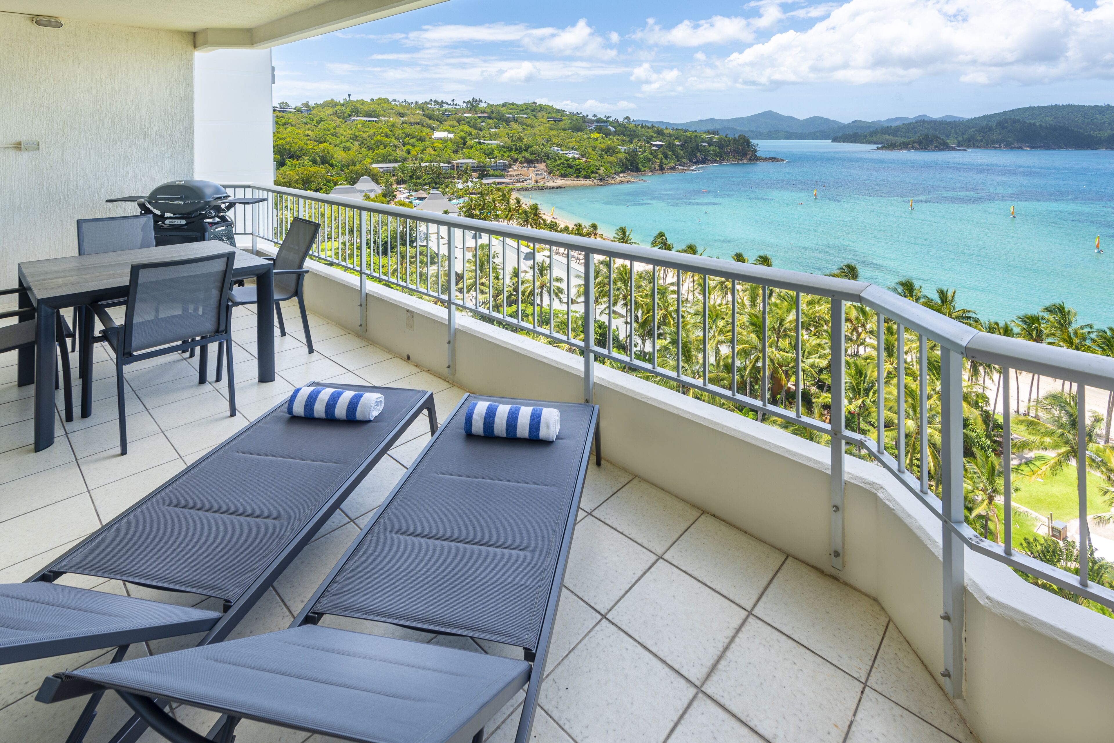 A balcony with two lounge chairs, a table, and chairs, overlooking a coastal view with clear skies.