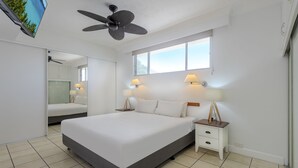 Un dormitorio con cama, mesitas de noche, lámparas, ventilador de techo, televisor y un ventanal grande.