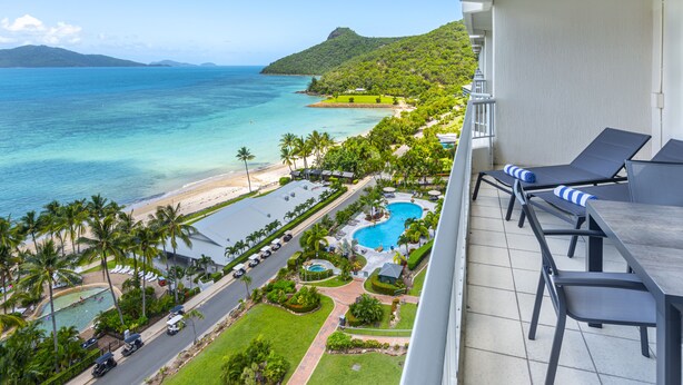 Un balcón de hotel con vista a una playa, una piscina y exuberante vegetación.
