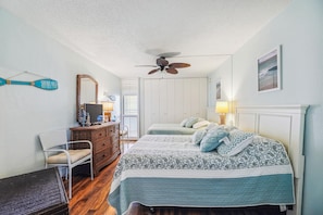 Une chambre avec deux lits, une commode, une chaise, un ventilateur de plafond et une fenêtre avec des stores.