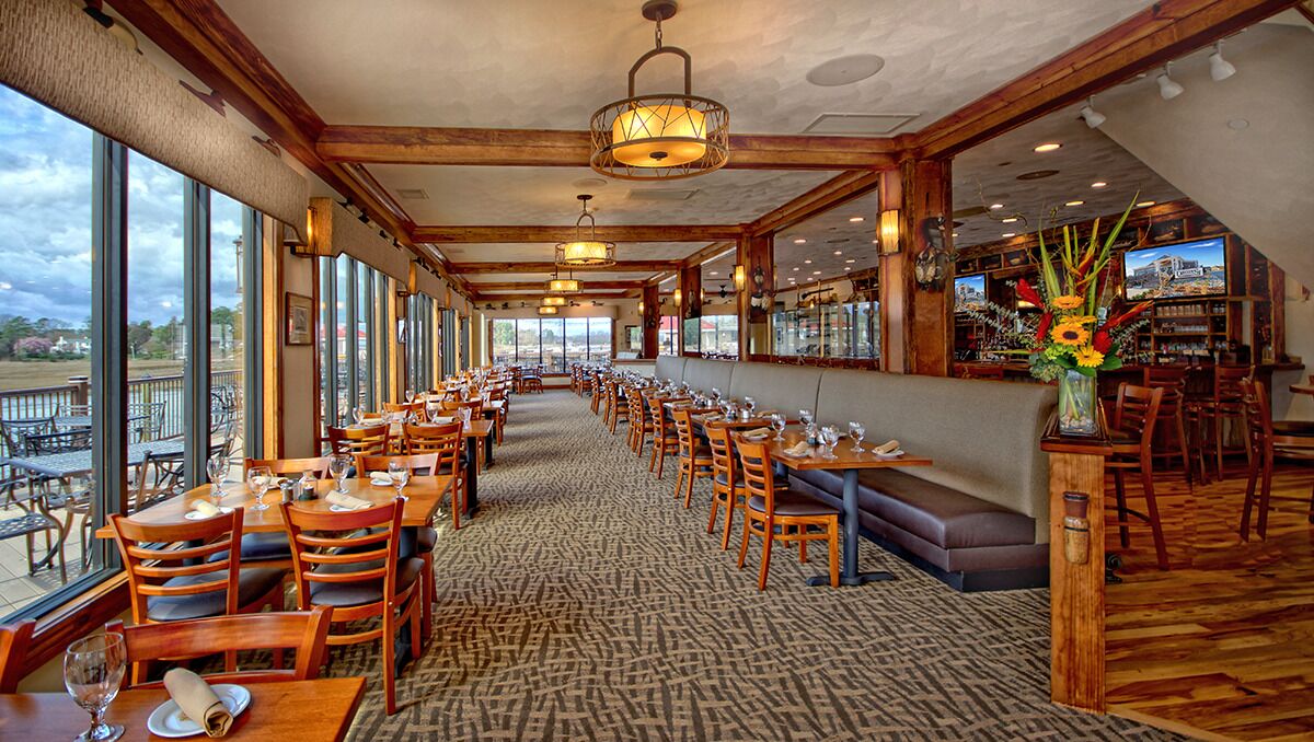 A restaurant with a long dining area, wooden tables and chairs, a bar area, and large windows offering a view.