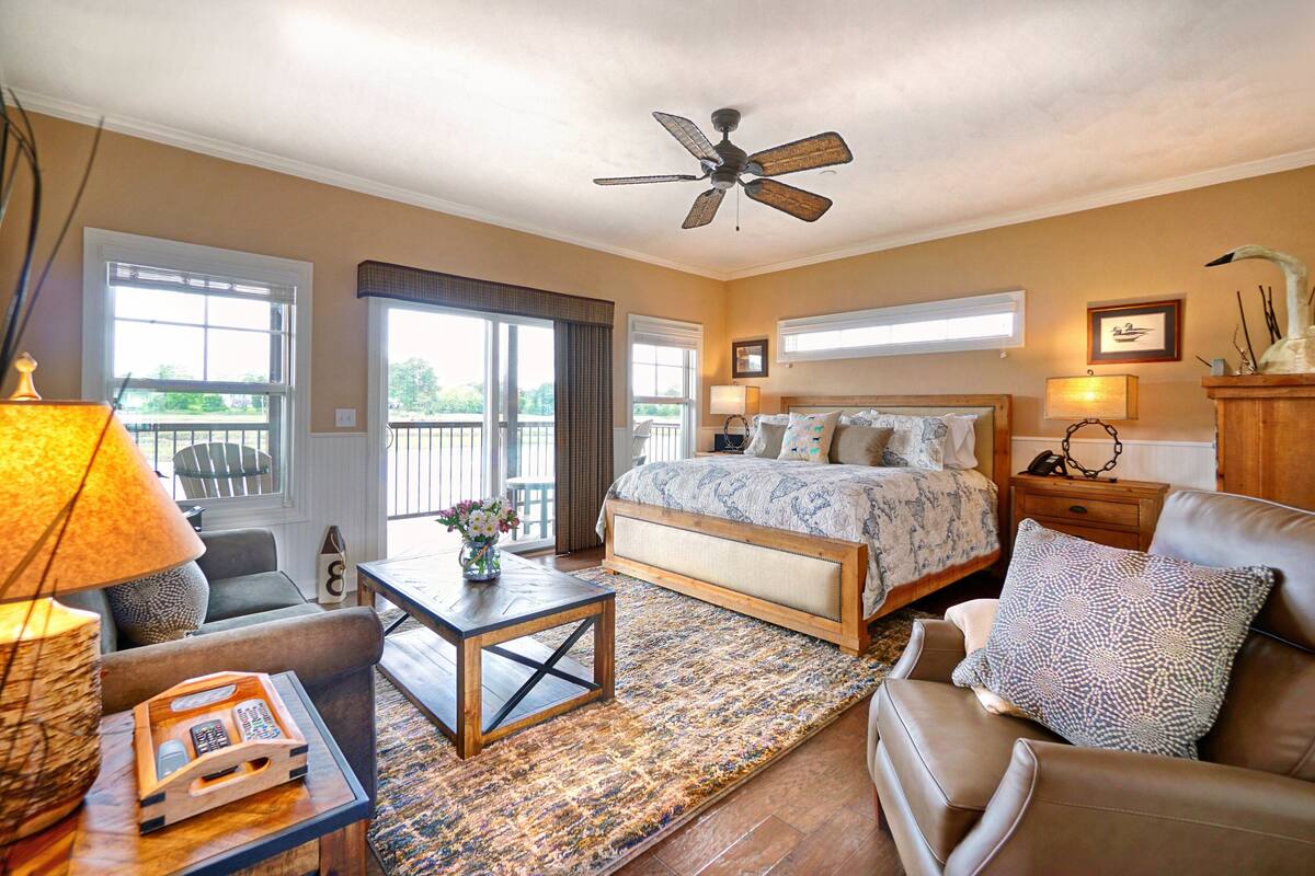 A bedroom with a bed, a ceiling fan, a nightstand, a lamp, and a view of the outdoors.