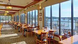 A restaurant with wooden tables and chairs, large windows with a view of the water, and a patterned carpet. - Decoys (Suffolk)