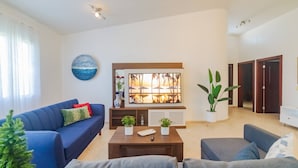 A modern living room with a blue sofa, a grey armchair, a coffee table, and a large window with a view of palm trees. - 7br Massive Villa In Sosúa - 2mins From Nightlife! (Sosúa)