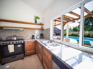 A modern kitchen with granite countertops, wooden cabinets, a stove, and a large window overlooking a pool. - Sun & Soul - Caribbean 9br Villa In Sosúa! (Sosúa)