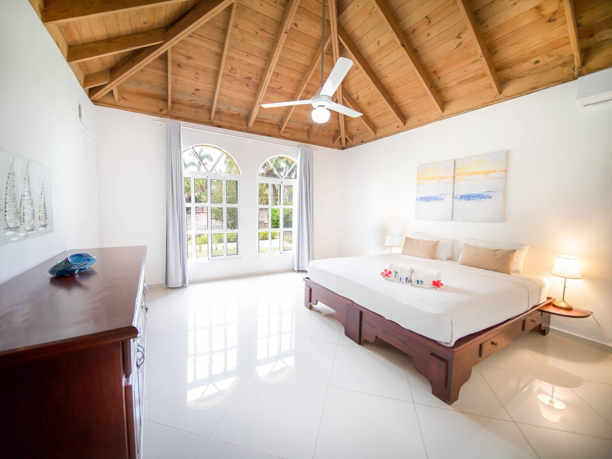 A hotel room with a wooden ceiling, a bed with a white bedspread, a nightstand with a lamp, and a view of the outdoors through a window.