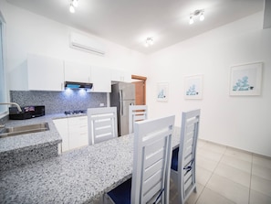 Een moderne keuken met witte kasten, een granieten aanrecht, een ingebouwde oven en een koelkast. Er staan witte stoelen bij een kleine eettafel.