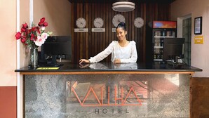 Un mostrador de recepción con un cartel que dice "RECEPTION" y "KAILA HOTEL" debajo. Hay relojes en la pared, un florero con flores y un ventilador de techo arriba.