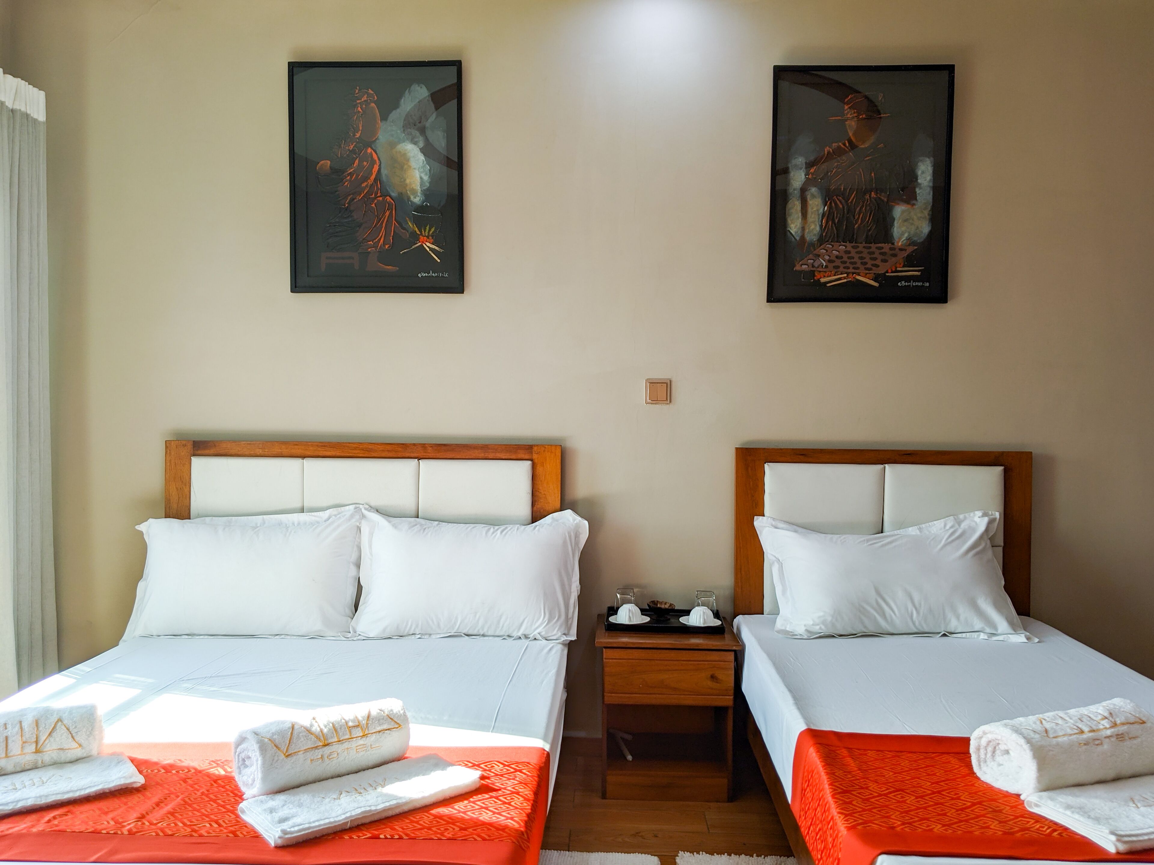 Une chambre d’hôtel avec deux lits, chacun agrémenté de draps blancs et d’une couvre-lit orange. Deux tableaux ornent les murs, il y a une table de chevet avec une lampe, et des serviettes sont posées sur les lits.