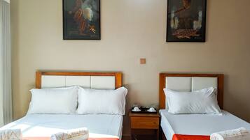Une chambre d’hôtel avec deux lits, chacun agrémenté de draps blancs et d’une couvre-lit orange. Deux tableaux ornent les murs, il y a une table de chevet avec une lampe, et des serviettes sont posées sur les lits.