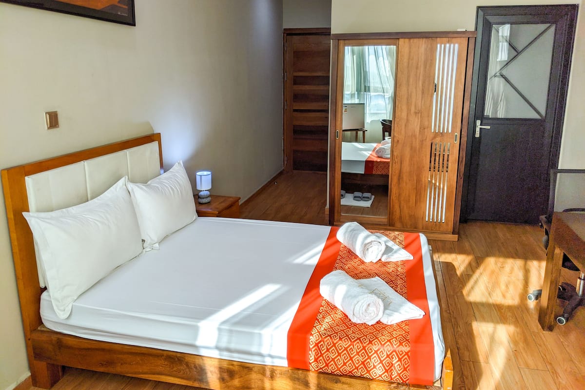 A hotel room with a wooden bed, a nightstand with a lamp, a bathroom with a shower, and a wardrobe.