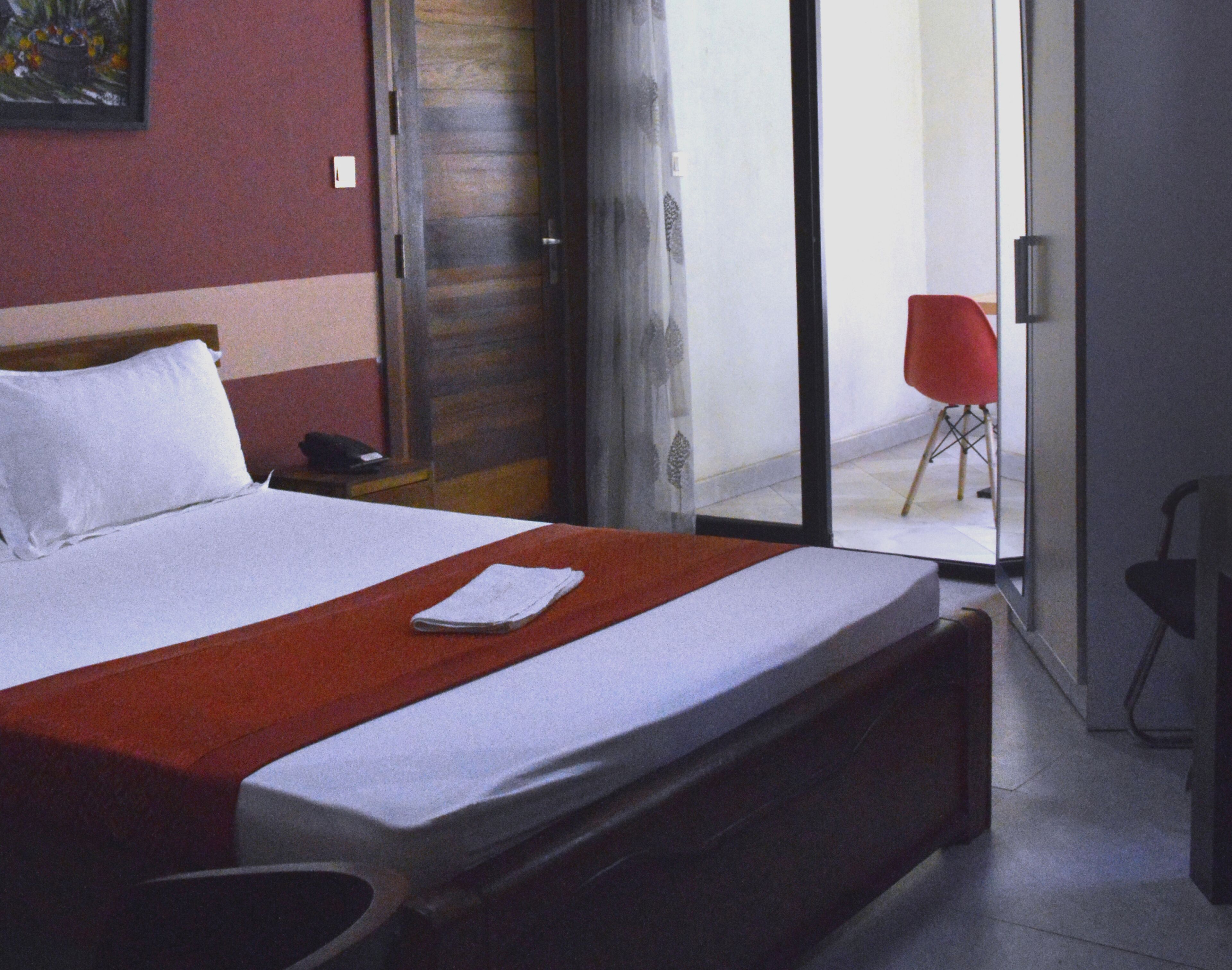 A hotel room with a bed, a red chair, a desk, and a sliding glass door leading to a balcony.