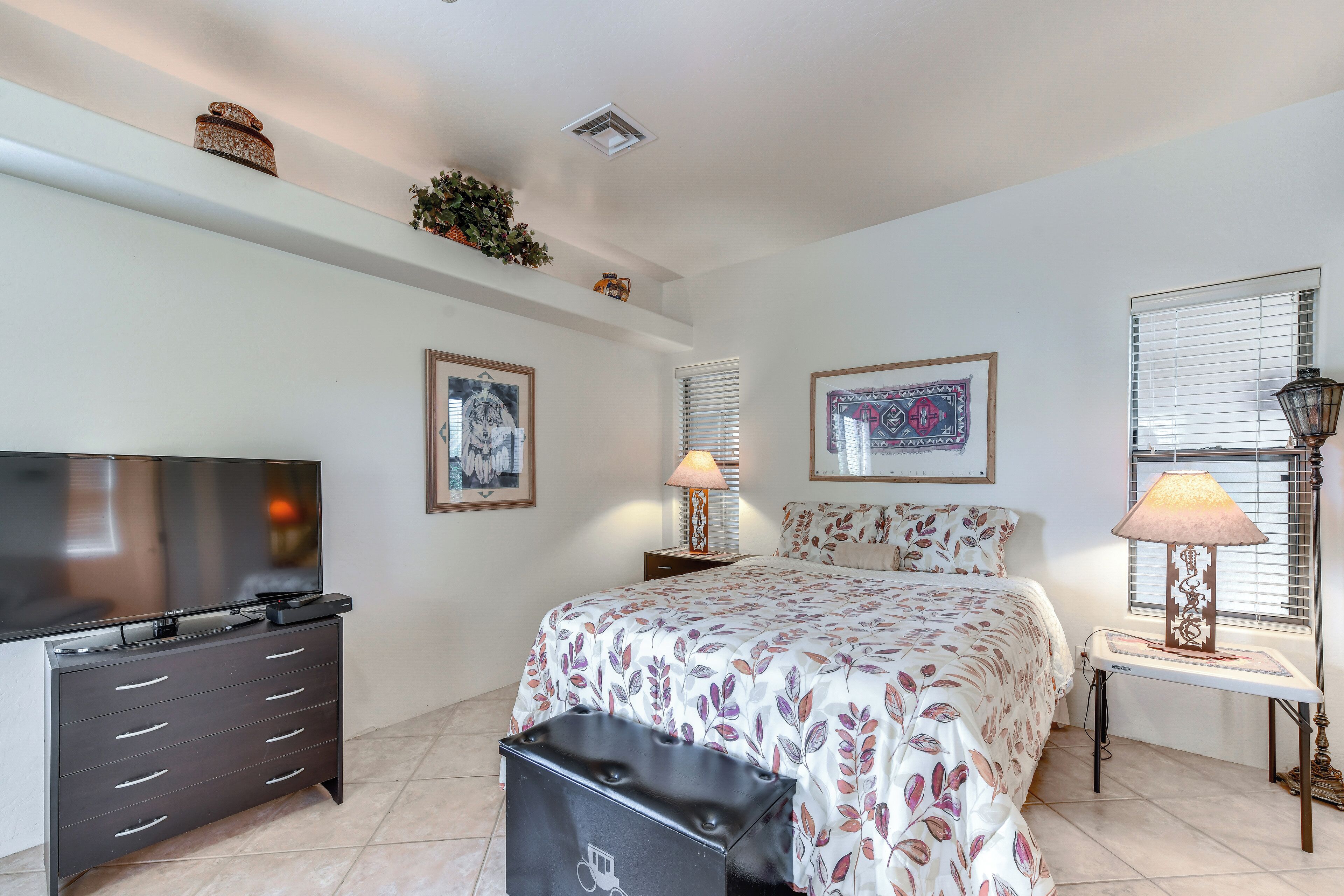 Un dormitorio con cama, televisor, cómoda, lámpara y obras de arte en las paredes.