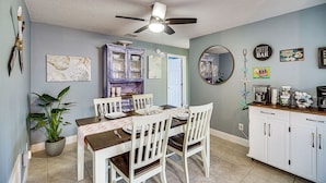Un coin repas avec une table dressée pour quatre personnes, des chaises blanches, un ventilateur de plafond et un espace cuisine équipé de placards et d’un coin pour préparer le café.