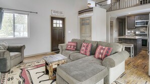 A spacious living room with a grey sofa, a wooden coffee table, and a patterned rug. A kitchen is visible in the background with modern appliances. - Hike + Ski: Grand Lake Cabin w/ Mtn-view Deck (Grand Lake)