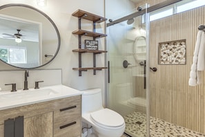 Un baño moderno con cabina de ducha de vidrio, inodoro, lavabo con espejo y estante de madera con un letrero que indica 'papel higiénico para descarga únicamente'.