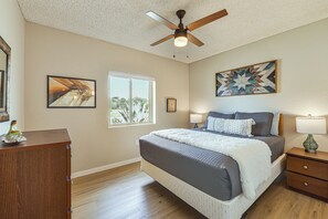 Un dormitorio con cama, mesitas de noche, ventilador de techo, ventana y obras de arte en las paredes.