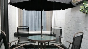 Uma mesa de vidro redonda com um guarda-sol preto, rodeada por quatro cadeiras com encosto em rede, posicionada diante de uma parede de tijolos brancos com uma janela ao fundo.
