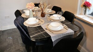 Mesa de jantar posta com pratos brancos, copos e talheres, rodeada por cadeiras pretas, com uma obra de arte emoldurada acima e uma janela com flores ao lado.