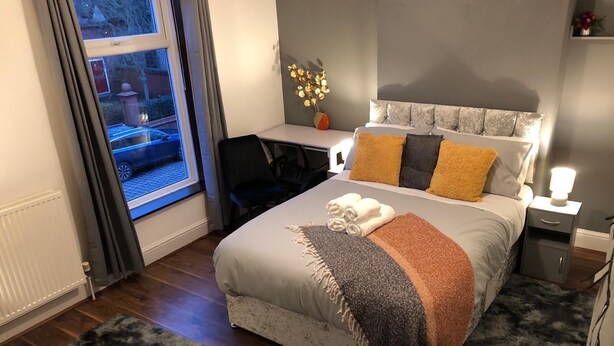 Quarto com cama, mesas de cabeceira, escrivaninha, cadeira, janela com cortinas e vista para o exterior.