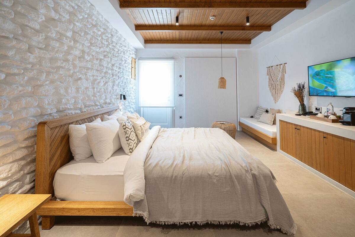 A bedroom with a stone wall, wooden ceiling, a bed with multiple pillows, a bedside table, a small round ottoman, a wooden bench, a TV on the wall, and a window with blinds.