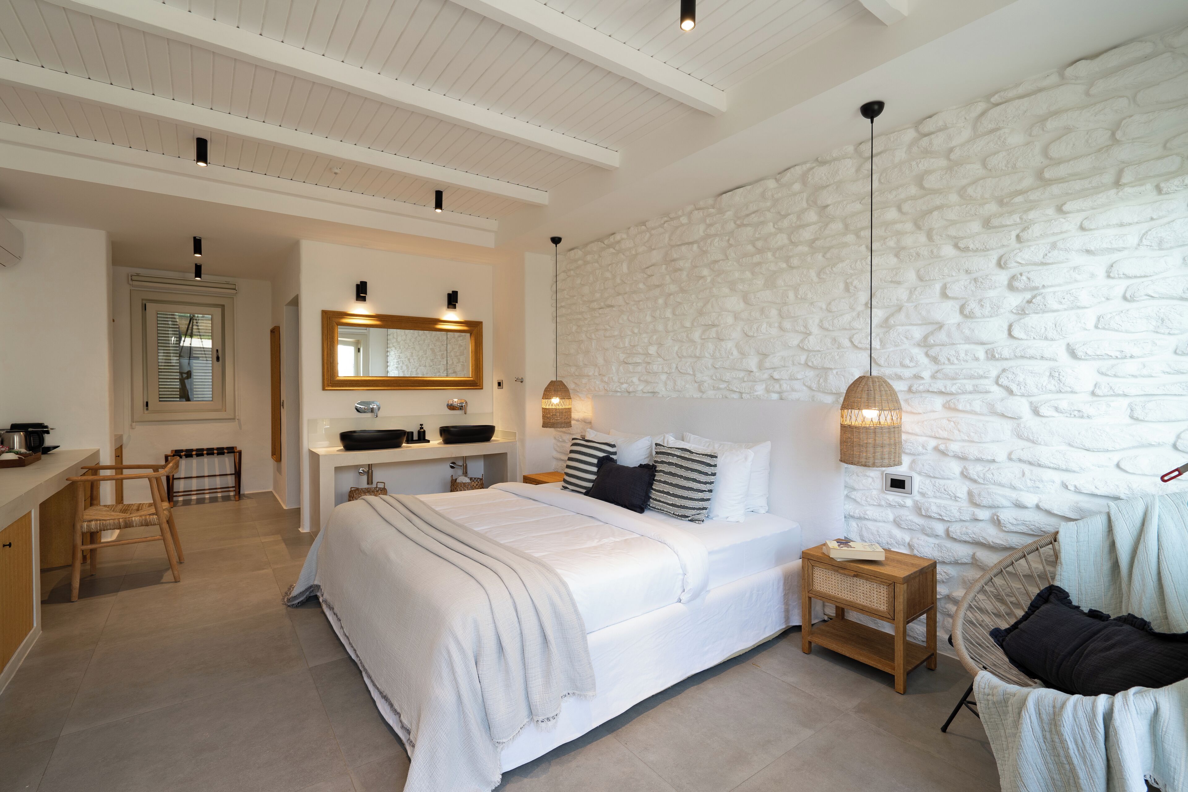 A bedroom with a large bed, a wooden desk, a chair, a mirror, and a stone wall.
