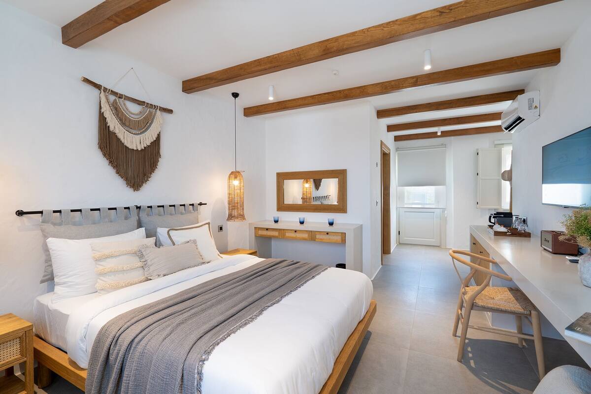 A modern bedroom with a large bed, wooden beams, a bedside table, a hanging macrame wall decoration, a bedside lamp, a mirror, and a TV.