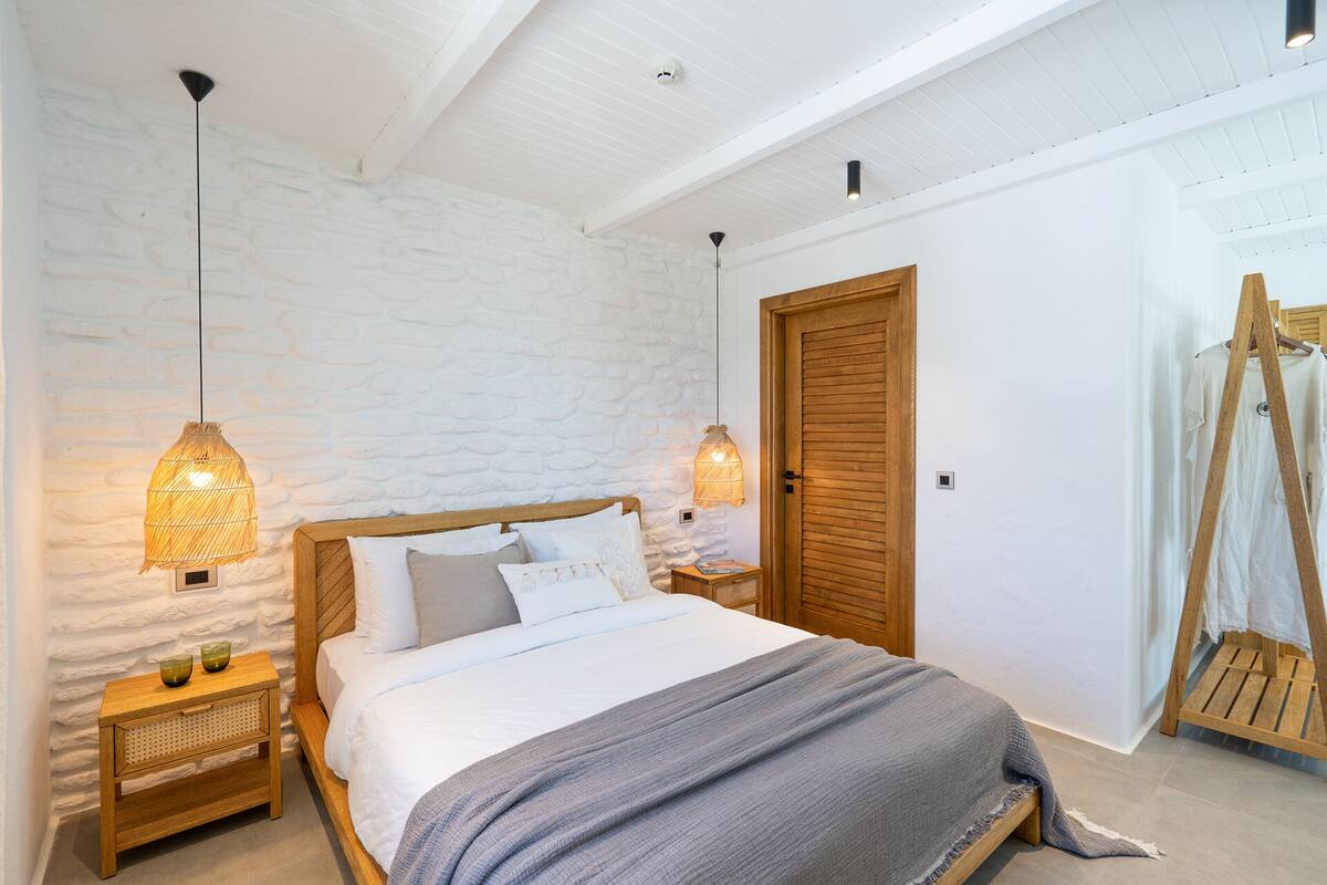 A bedroom with a wooden bed, white walls, and a stone-like textured wall. There are two bedside tables with lamps, a wooden wardrobe, and a wooden ladder.
