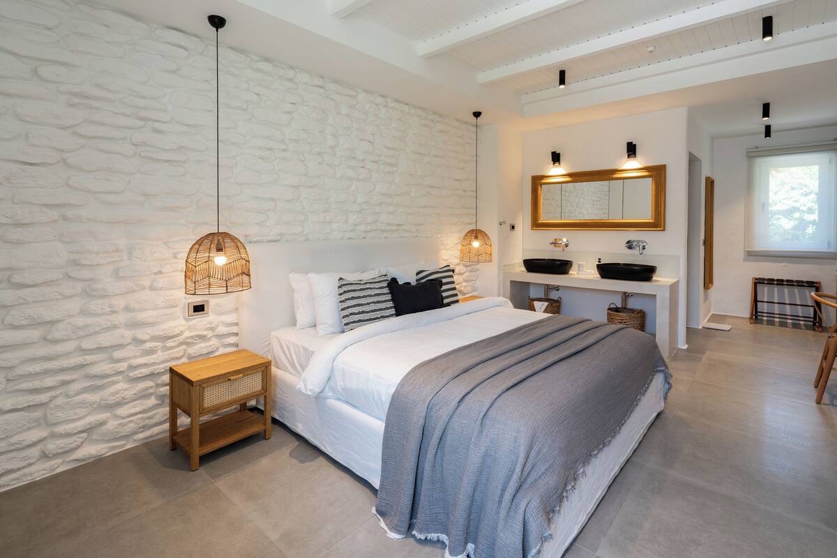 A bedroom with a stone wall, a bed with a grey blanket, bedside lamps, a wooden nightstand, a large mirror, and a window with a view.