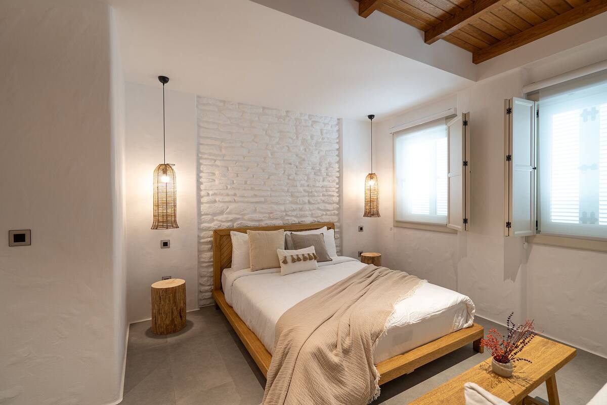 A bedroom with a bed, two bedside tables, a wooden headboard, a white brick wall, and a window with blinds.