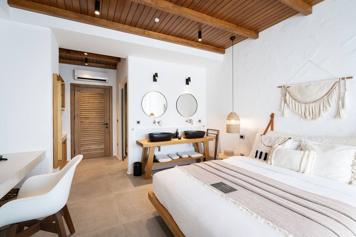 A modern bedroom with a large bed, a wooden desk, two round mirrors, a bedside table with two black bowls, and a hanging macrame piece.