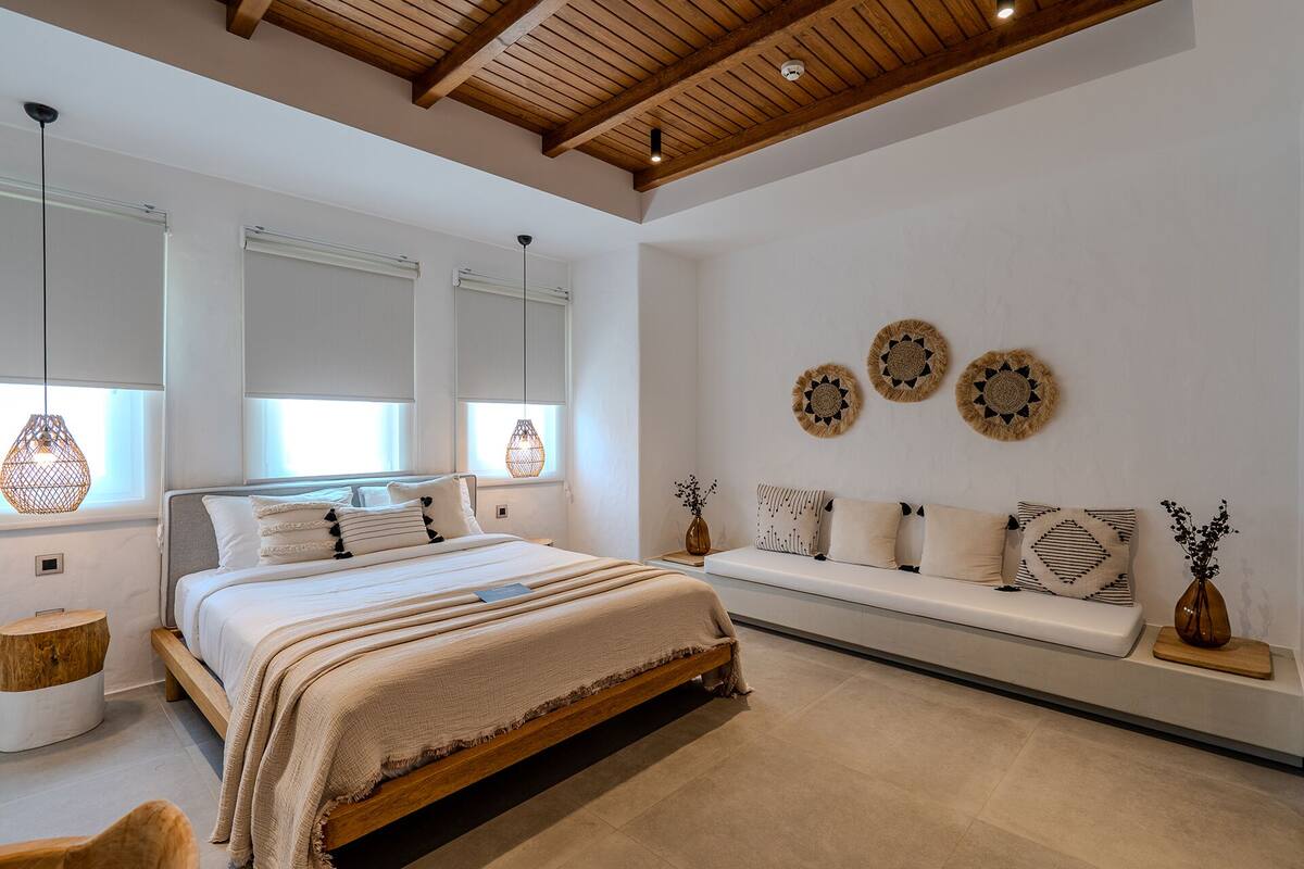 A modern bedroom with a wooden ceiling, a large bed with a wooden frame, a bench with cushions, and decorative wall art.