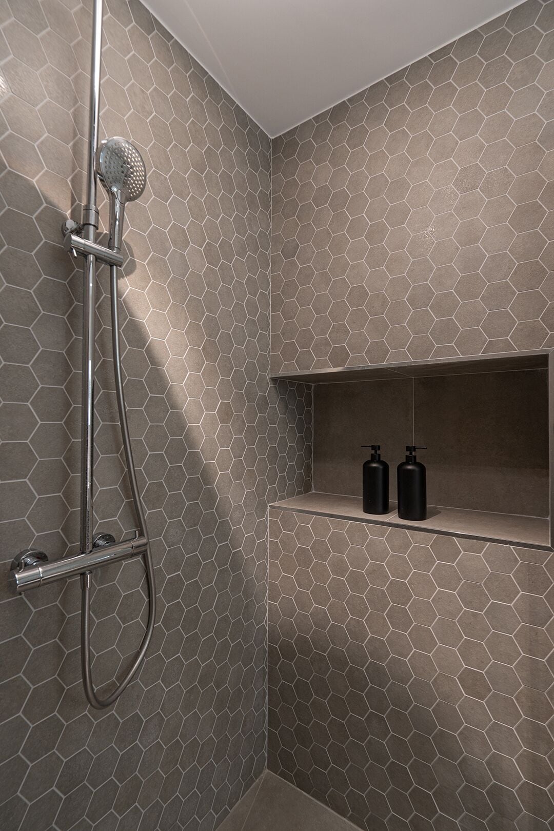Una ducha con un patrón de azulejos hexagonales y un estante con dos botellas negras.
