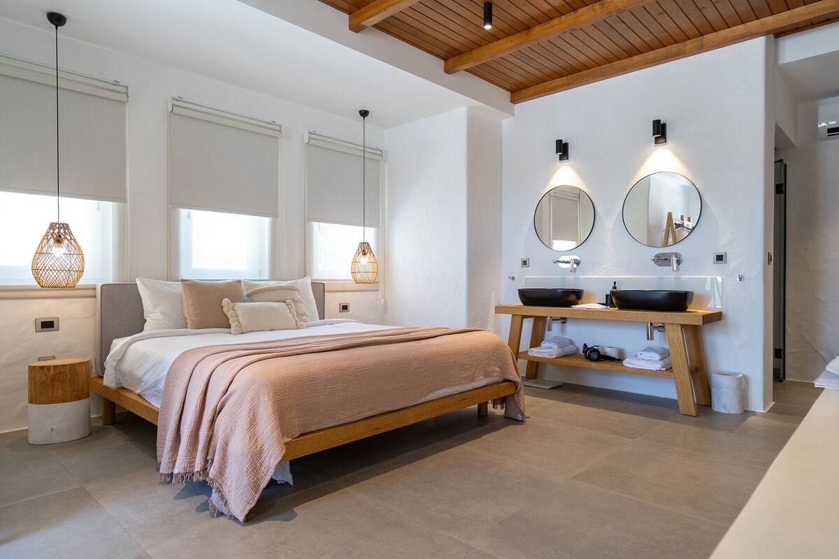 A modern bedroom with a large bed, bedside tables, a wooden headboard, a built-in wooden shelf with towels, a round mirror, and a large window with blinds.