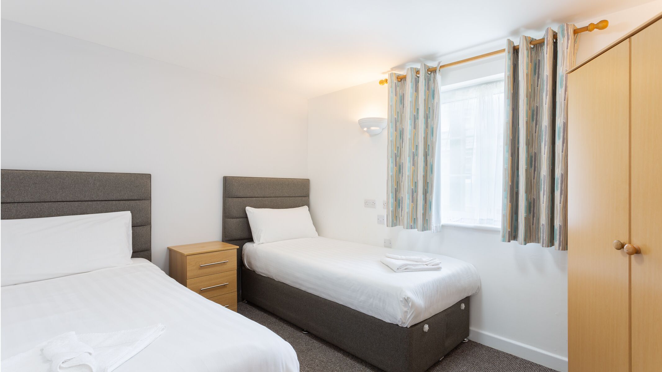 Une chambre avec deux lits, une table de chevet, une armoire, une fenêtre avec des rideaux et un luminaire fixé au mur.