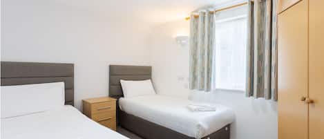 Une chambre avec deux lits, une table de chevet, une armoire, une fenêtre avec des rideaux et un luminaire fixé au mur.